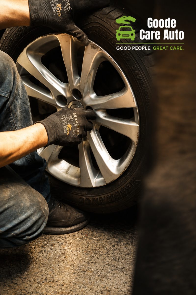 A mechanic wearing gloves is adjusting a car wheel and tire beside the Goode Care Auto logo.